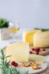 Piece of cheese, cheese wheel, rosemary greens and glass of wine in background, selective focus