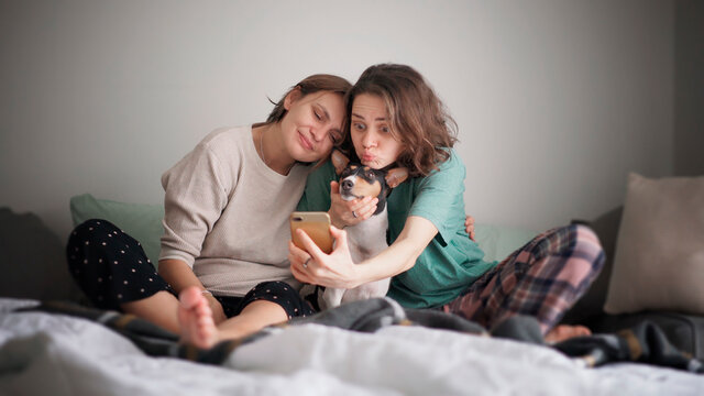 Beautiful happy lesbian couple sitting in pajamas on the bed in the morning making selfie on smartphone with dog bassengi - Powered by Adobe