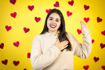 Young caucasian woman over yellow background with red hearts smiling swearing with hand on chest...