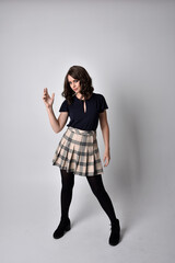 Full length portrait of pretty brunette woman wearing tartan skirt and blouse.  Standing pose on the ground,  against a  studio background.