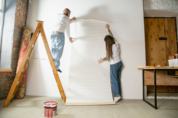 Young couple, family doing apartment repair together themselves. Married man and woman doing home makeover or renovation. Concept of relations, moving, love. Hanging wallpaper with ladder.
