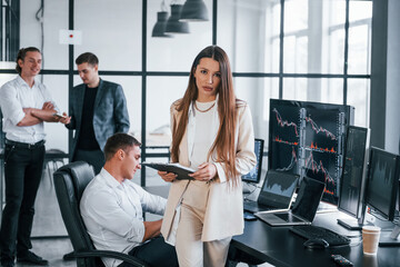 Young woman in formal wear standing of front of people. Team of stockbrokers works in modern office with many display screens