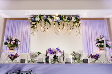 Festive table, arch, stands decorated with composition of violet, purple, pink flowers and greenery, candles in the banquet hall. Table newlyweds in the banquet area on wedding party.