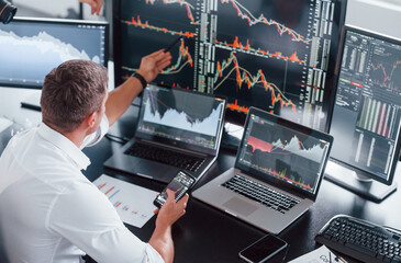 In formal clothes. Young male stock brocker works indoors in the office. Many of displays