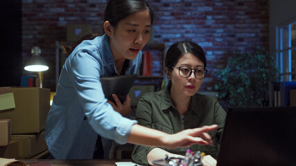 Two young Asian women startup small business entrepreneur SME distribution warehouse with parcel in night. chinese lady colleagues working together on laptop computer discussing to solve problem.
