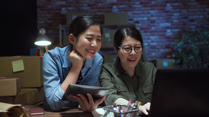 Young happy asian business women work together by using laptop computer and touchpad with parcel boxes packaging at their startup home office in dark night. SME online business seller and delivery.