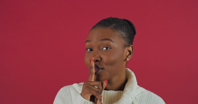 Portrait of afro american woman gossip girl ethnic girl showing silence gesture puts finger on mouth forbidding to speak make noise looks at camera stands on red background in studio, mystery concept