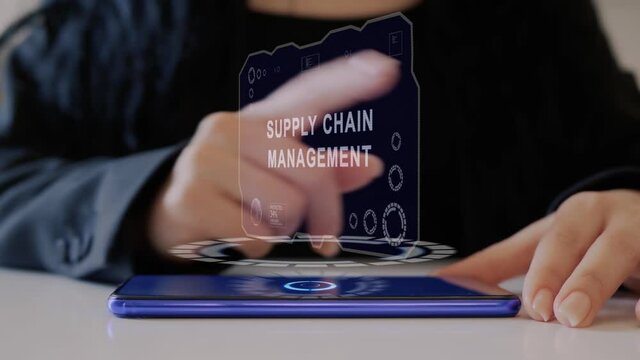 Female Hand Interacts HUD Hologram With Text Supply Chain Management. Woman In Black Uses Conceptual Holographic Technology Of The Future On The Screen Of A Smartphone Lying On A Table