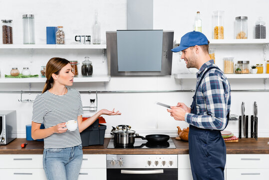 young woman with cup of coffee talking to handyman with tablet in kitchen - Powered by Adobe