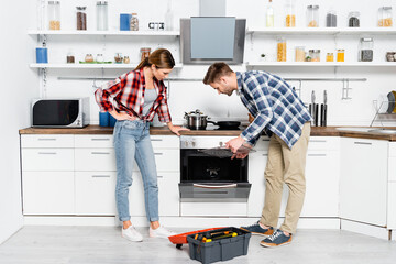 full length of young couple bending over disassembled oven and open toolkit in kitchen