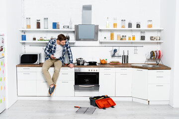 full length of young man with tablet looking at disassembled oven while sitting on table in kitchen