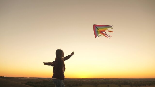 A happy girl runs with a kite in her hands across field in rays of the sunset. The child wants to be pilot. A healthy child dreams of freedom, flight. Kid plays outdoors in the park.