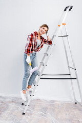 Fototapeta na wymiar full length of happy young woman with paint roller looking at camera while leaning on ladder at home