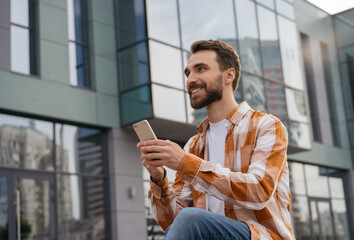 Young man using mobile phone, working online, sitting outdoors. Successful freelancer holding smartphone, receive payment