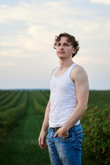 Young handsome man, wearing torn jeans and white t-shirt, standing on green field, thinking. Artistic education concept. Creative male portrait outside.