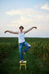 Fototapeta premium Young handsome man, wearing torn jeans and white t-shirt, sitting on yellow stool on green field, thinking. Artistic education concept. Creative portrait outside.
