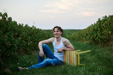 Young handsome man, wearing torn jeans and white t-shirt, sitting by yellow stool on green field,...