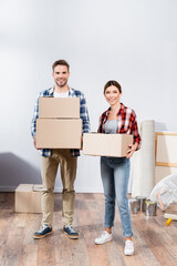 Fototapeta na wymiar full length of happy young couple looking at camera while holding cardboard boxes at home