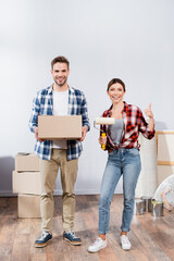 full length of happy young woman with paint roller showing thumb up near man with cardboard box at home