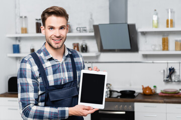 front view of happy handyman looking at camera while showing tablet with blurred kitchen on background