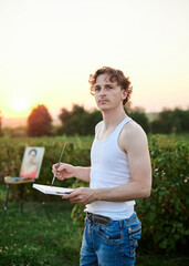 Young male artist, wearing torn jeans and white t-shirt, standing on green field during sunset, holding empty canvas, thinking. Painting workshop in countryside. Artistic education concept.