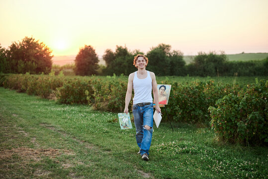 Young male artist, wearing torn jeans and white t-shirt, walking on green field during sunset, holding canvas and palette thinking. Painting workshop in countryside. Artistic education concept.