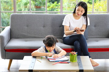 Asian mother shopping online from home using mobile phone while watching her daughter working on art class assignment from school for homeschooling and education concept