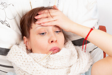 a young pretty girl is sick at home in bed, holds her hot forehead and measures the temperature with a thermometer