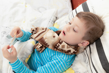 Obraz premium a sick child a boy in a blue jacket lies at home in bed and measures the temperature with a thermometer