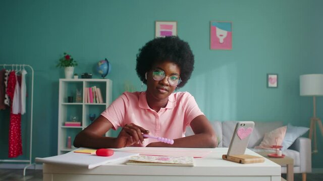 Portrait of young woman, stylish student in pink T-shirt and cool glasses, sitting at desk at home and playing with purple pencil, looking at camera and smiling, Zoom out, Slow motion.