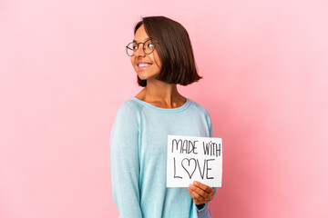 Young hispanic mixed race woman holding a made with love paper poster looks aside smiling, cheerful and pleasant.