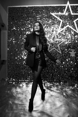 Sexy brunette woman with long wavy hair in black blazer, net body, short leather shorts, tights and shoes standing and posing against shining silver background with stars. Full length portrait