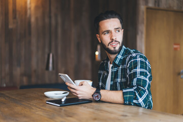 Pensive hipster guy with modern cellphone device thoughtful looking away while creating publication...