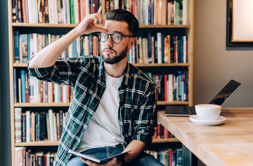 Contemplative male student in optical eyewear holding education textbook thinking about informative...