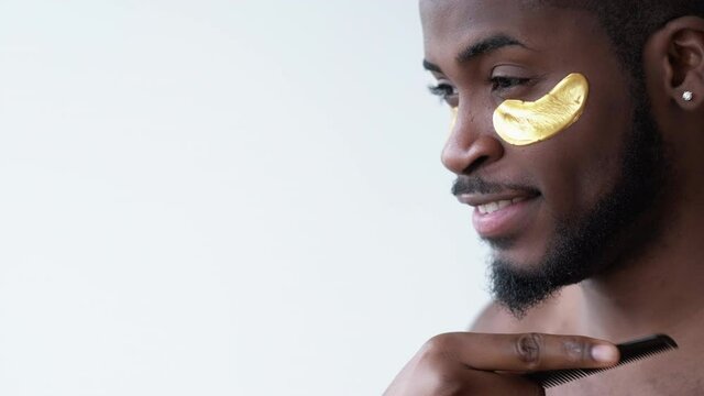 Man grooming. Facial treatment. Metrosexual lifestyle. Profile of confident satisfied African guy with gold collagen gel eye patches combing beard smiling isolated on neutral background.
