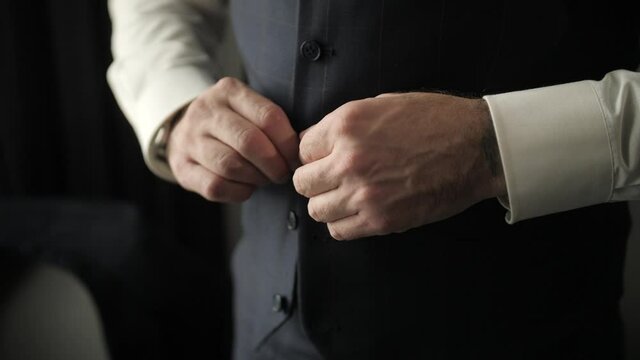 The man at the window dresses in a stylish suit and tidies himself up. The groom fastens a button on his vest. Preparing for the event. The future groom dresses up before meeting the bride.