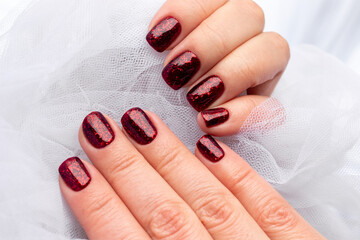 Female hands with beautiful manicure - dark red glittered nails in white tulle fabric. Selective focus