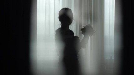 Silhouette of a girl at the window in a dark key. Wedding photo session in the room at dawn. A beautiful bride in a peignoir poses by the window for photo and video shooting. Morning of the bride.