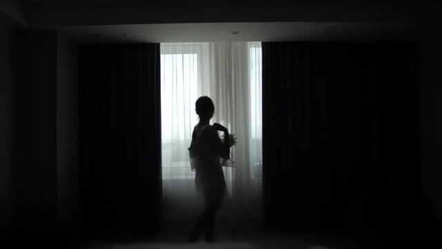 Silhouette of a girl at the window in a dark key. Wedding photo session in the room at dawn. A beautiful bride in a peignoir poses by the window for photo and video shooting. Morning of the bride.