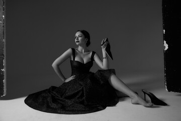 Elegant sexy woman in black evening shiny dress sitting on white floor in studio and posing, holding her shoe in hand. Sexy girl with perfect makeup and brunette hair in bun