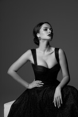 Elegant woman with perfect make-up and brunette hair in a bun in black shiny evening ball gown dress with sexy decolette sitting at white cube against grey background. Studio portrait