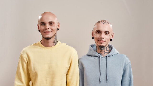Beat friends. Portrait of young happy caucasian twin brothers with tattoos and piercings smiling at camera while posing together in studio, standing isolated over beige background