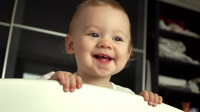 Close up of little baby looks at camera, smiles and laughs after sleeping in crib. Happy half year old girl smiles and shows first teeth.