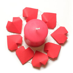 Thick pink aromatic candle on white openwork paper napkin, surrounded by a several 3D handmade paper hearts on white background top closeup view isolated. Love, rendezvous, relations, dating concept