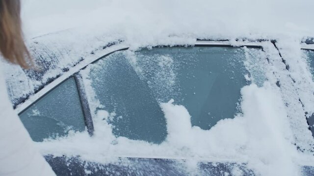 Female hand with brush in hand sweeping snow from side window of black car in winter season