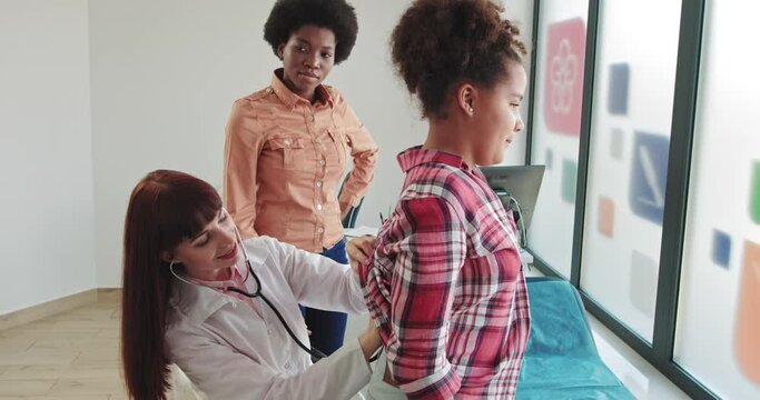 Young Woman In A White Coat, Pediatrician Examines A Sick Girl, Listens To Breathing Using A Phonendoscope. Doctor And Child. Ambulance. Medium Shot. European Doctor And African Patient