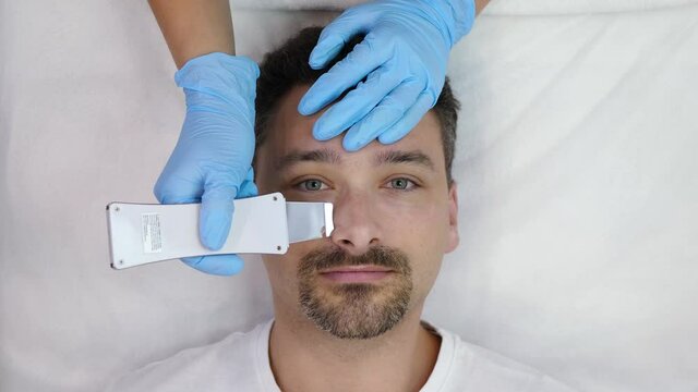 Bearded handsome middle aged man receives cosmetological ultrasonic facial cleaning, looking at camera and isolated on white background, top view. Beautician doing ultrasound peeling on male face
