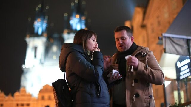 A Joyful Couple Eats Sweet Pastries In The Night City Against The Background Of Old Towers. A Man Smears A Woman's Nose. The Playful Mood Of Lovers On A Date.