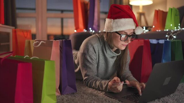 Happy woman with glasses wearing a santa claus hat is lying on the carpet and makes an online purchase using a credit card and laptop. Shopping bags around.