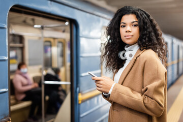 Obraz premium smiling african american woman in wireless headphones holding smartphone in subway